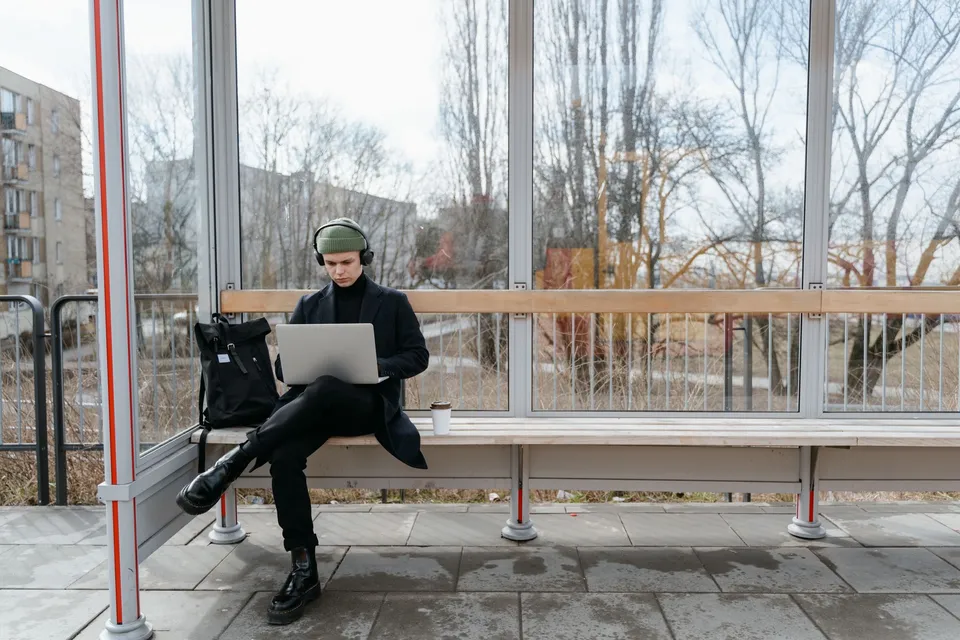 A young man works remotely on a laptop while sitting at a bus stop, showcasing modern digital nomad life.